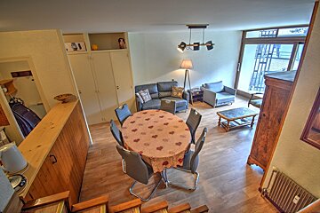 Overhead view of a living/dining room. Features a round table with a heart-patterned cloth, sofa, armchair, and large window. Wooden floors and warm lighting.