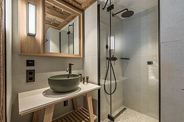 A chic modern bathroom with a light wood vanity, green bowl sink, and glass shower. Features concrete walls, wooden ceiling, and contemporary fixtures.