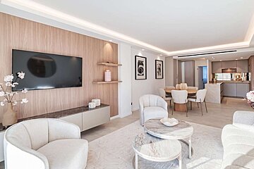 Bright, modern open-concept living room with light wood walls, TV, white furniture, marble tables, dining area, and kitchen in the background.
