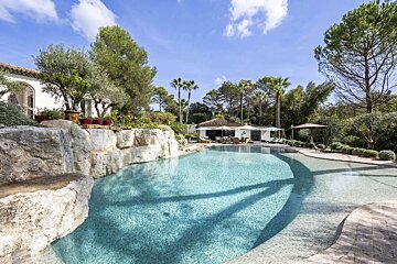 A sunny, luxurious outdoor pool with rock landscaping, surrounded by lush trees, and white buildings in the background under a blue sky.