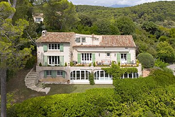 A charming Mediterranean villa with green shutters and terracotta roofs, nestled in a lush, forested hillside.