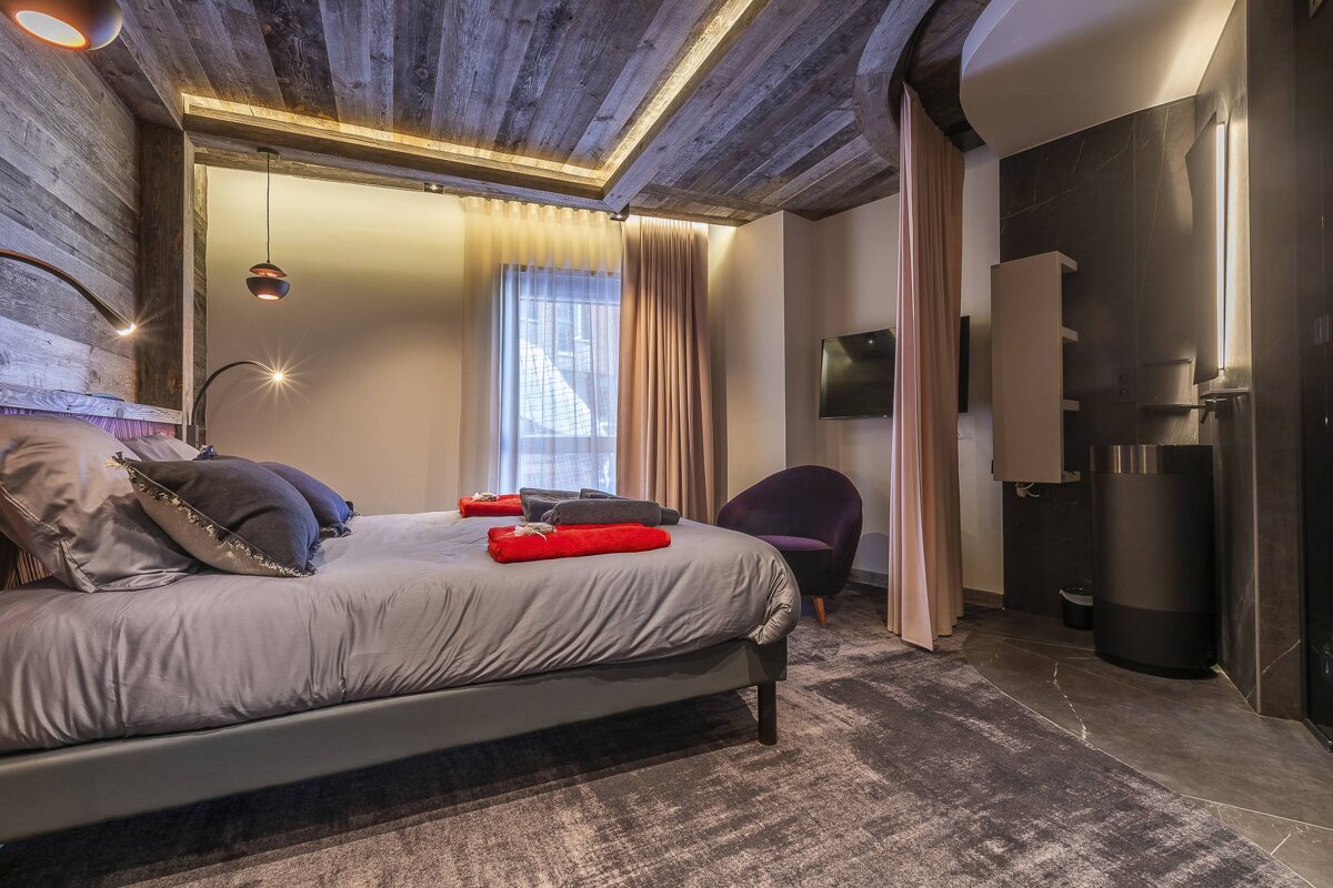A cozy modern bedroom featuring rustic wood ceilings, a grey bed with red accents, and warm lighting.