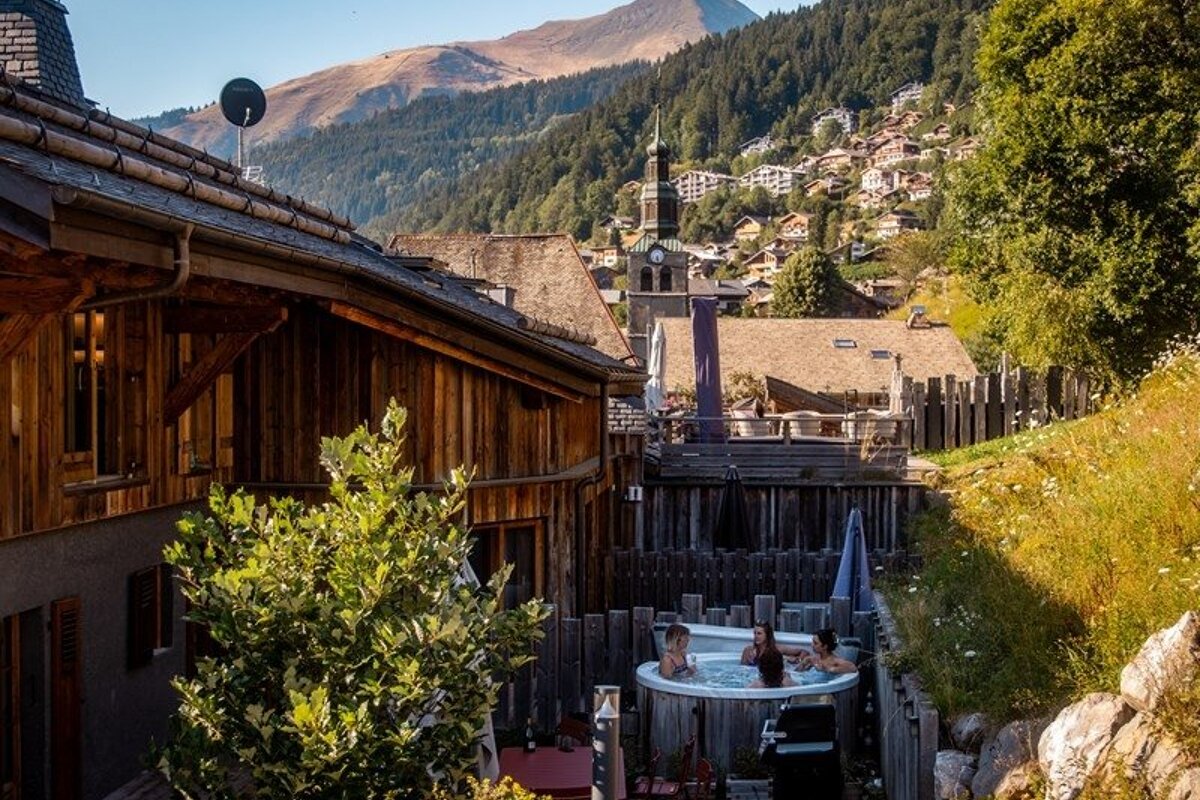 People relax in an outdoor hot tub next to a wooden chalet in a scenic alpine mountain village.