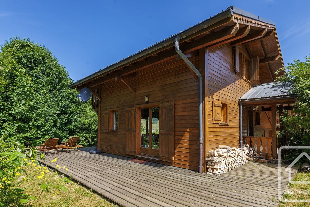 A charming wooden cabin with a wide wooden deck, nestled in a lush green setting under a clear blue sky, with stacked firewood nearby.