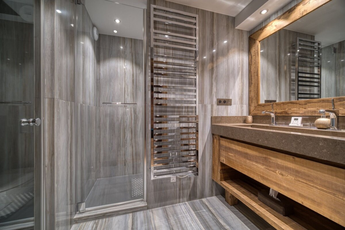 Elegant gray-tiled bathroom with a glass shower, chrome towel warmer, and a rustic wooden double vanity featuring a large mirror.