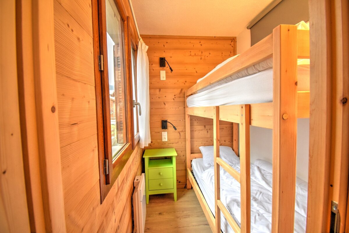 A cozy, wood-paneled room featuring bunk beds with white sheets, a window with curtains, and a bright green nightstand.