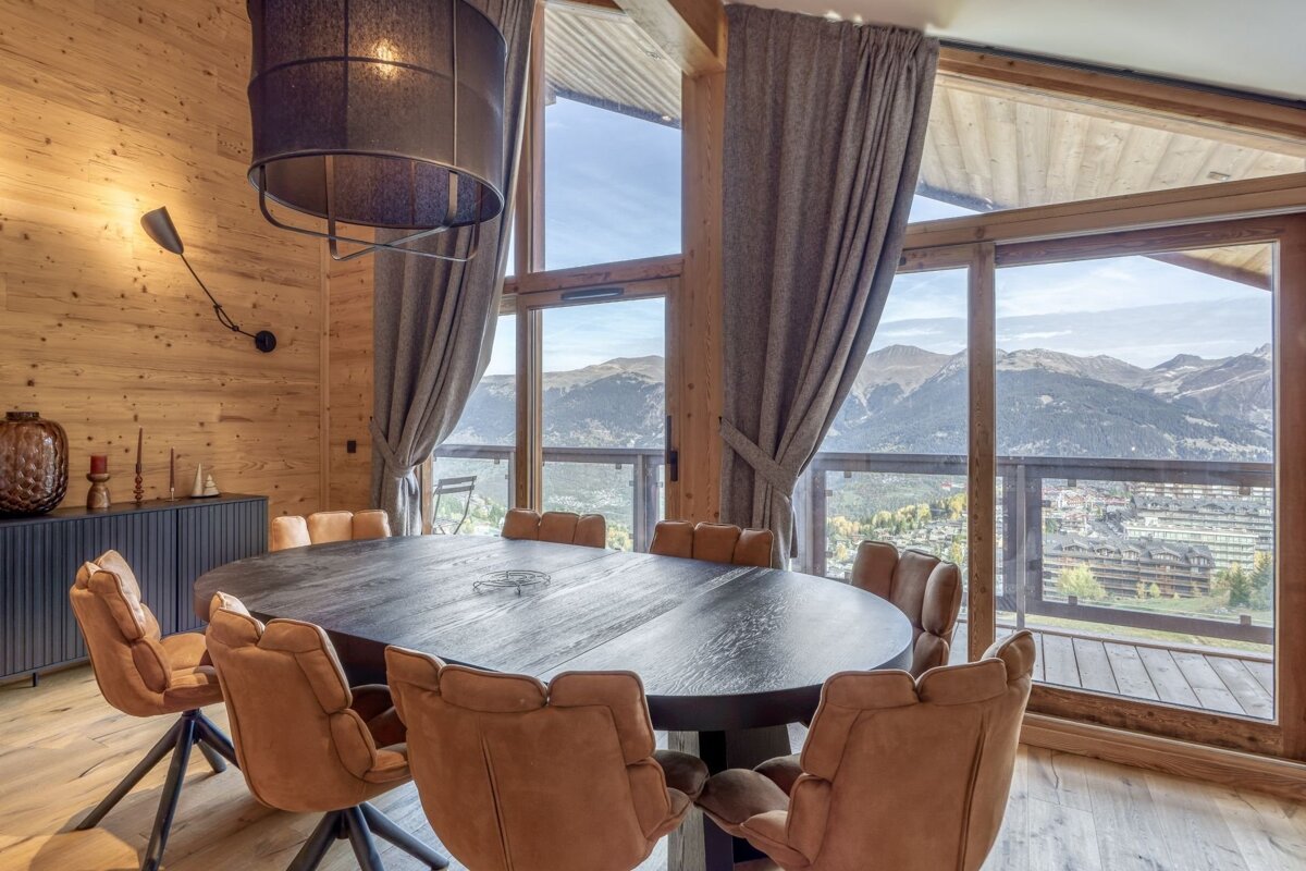 A cozy chalet dining room with panoramic mountain views through large windows. Features wooden walls, a dark oval table, and tan chairs.
