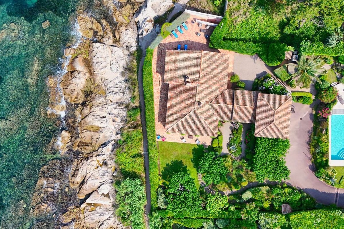 Aerial view of a coastal villa with a tiled roof, lush green gardens, a swimming pool, and a rocky shoreline with clear blue water.