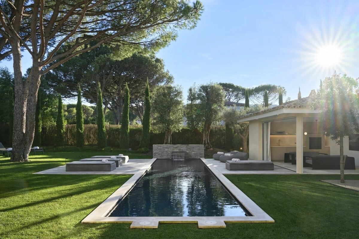 A luxurious outdoor living space with a dark rectangular pool, manicured green lawn, modern lounge areas, and a pool house under a bright, sunny sky.