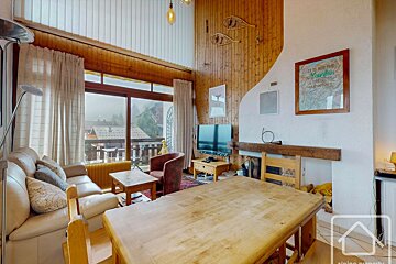 A rustic alpine living room with wood panels, a fireplace, large windows showing a foggy mountain view, and a large wooden dining table.