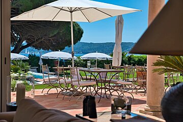A sunny patio with dining tables and umbrellas overlooking a blue sea with hills, viewed from a cozy indoor setting with a lounge and lamp.