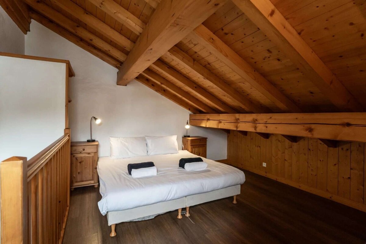 A rustic loft bedroom featuring a white bed, wooden nightstands, and prominent exposed timber beams on the angled ceiling.