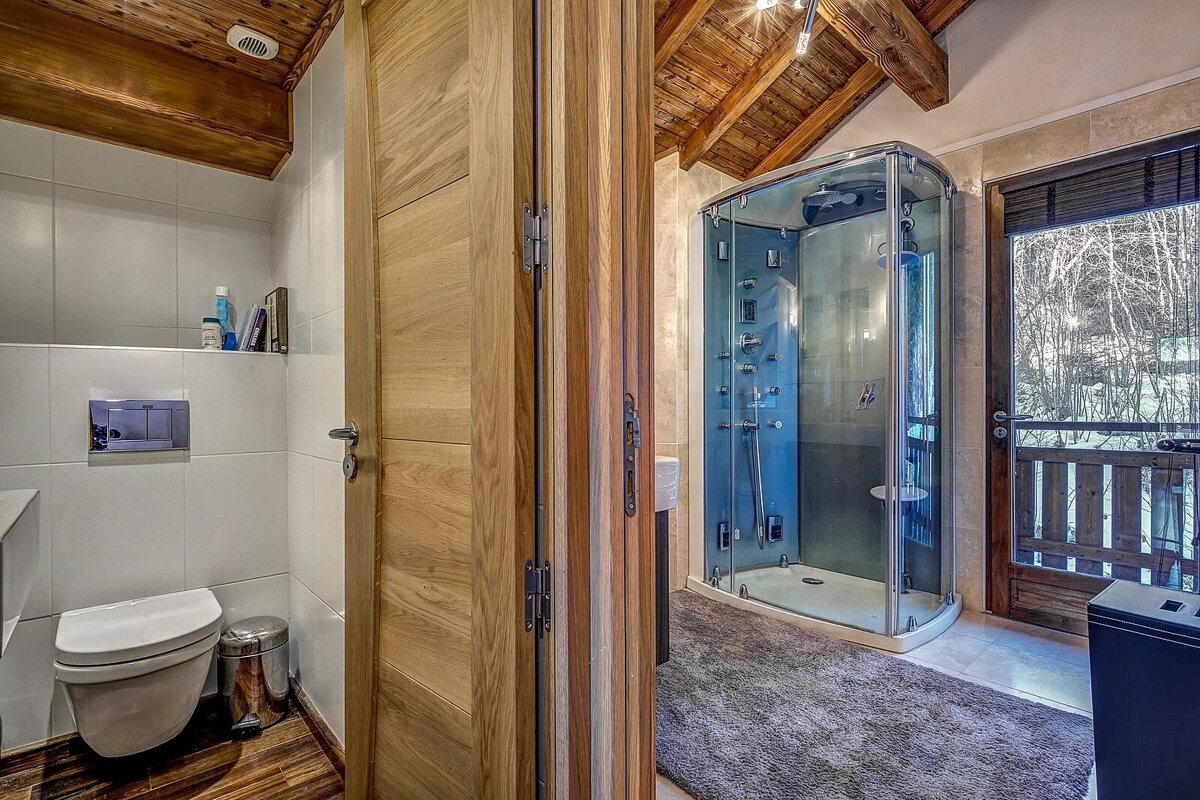 Rustic bathroom with exposed wooden beams, a modern glass shower cabin, toilet, and a folding door. A balcony overlooks a snowy forest.