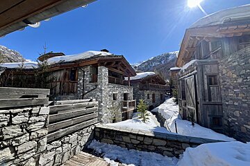 Snow-covered stone and wood chalets line a sun-drenched mountain village street, with bright blue skies and snowy peaks in the background.