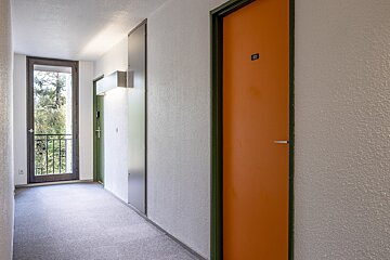 A bright hallway with white textured walls, grey carpet, an orange door (303), a green door, and a balcony overlooking trees.