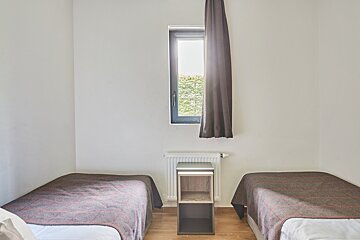 A minimalist twin bedroom with white walls, wooden floor, a window with a dark curtain, a radiator, and a small table between beds.