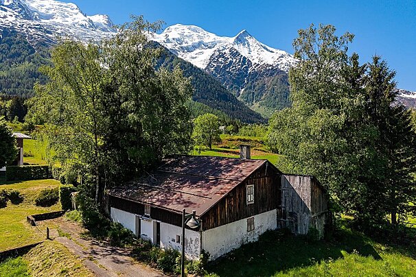 Rustic wooden cabin in a lush green valley with majestic, snow-capped alpine peaks rising in the background under a clear blue sky.
