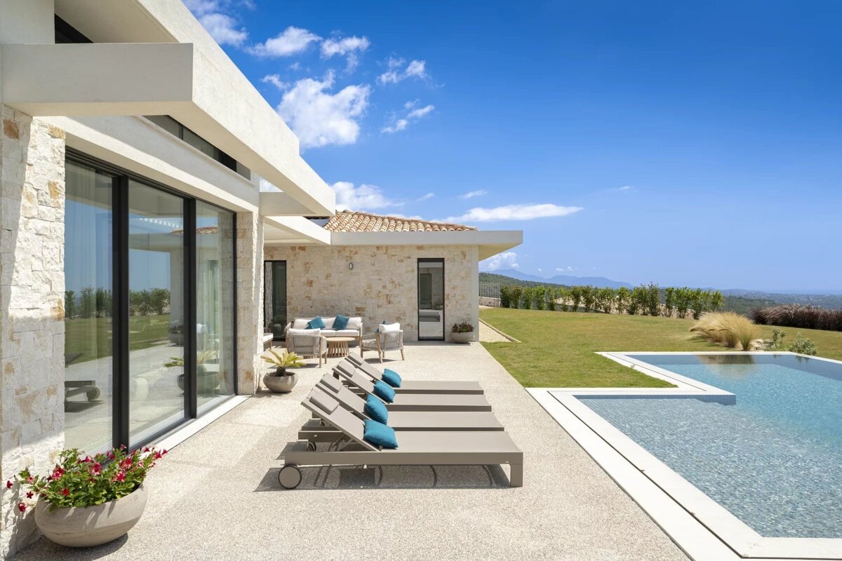 Modern stone villa with sun loungers beside a swimming pool, overlooking a green lawn under a bright blue sky.