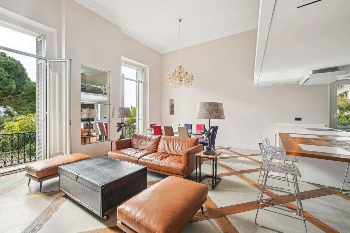 A bright, luxurious open-plan living space features brown leather furniture, a patterned floor, high ceilings, a chandelier, and a balcony with greenery.