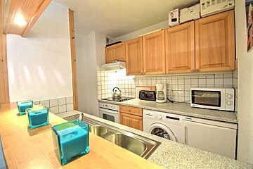 A small, well-equipped kitchen with light wood cabinets, white tiled backsplash, various appliances, and a counter with blue candles.