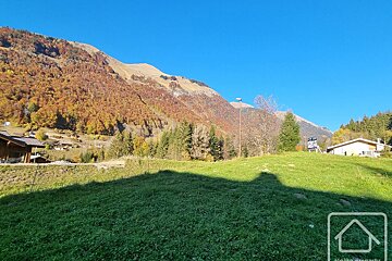 A sunny green field with mountains covered in vibrant autumn foliage under a clear blue sky.