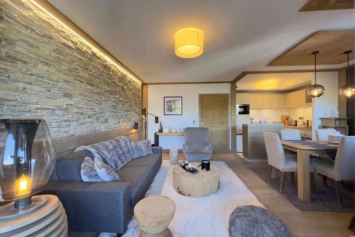 A cozy, open-plan living room, dining area, and kitchen with a rustic stone accent wall, grey sofa, wooden furniture, and warm ambient lighting.