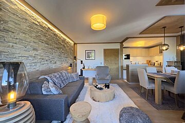 A modern rustic open-plan living/dining/kitchen space. Features a stone accent wall, grey sofa, wooden furniture, and warm lighting on a white rug.