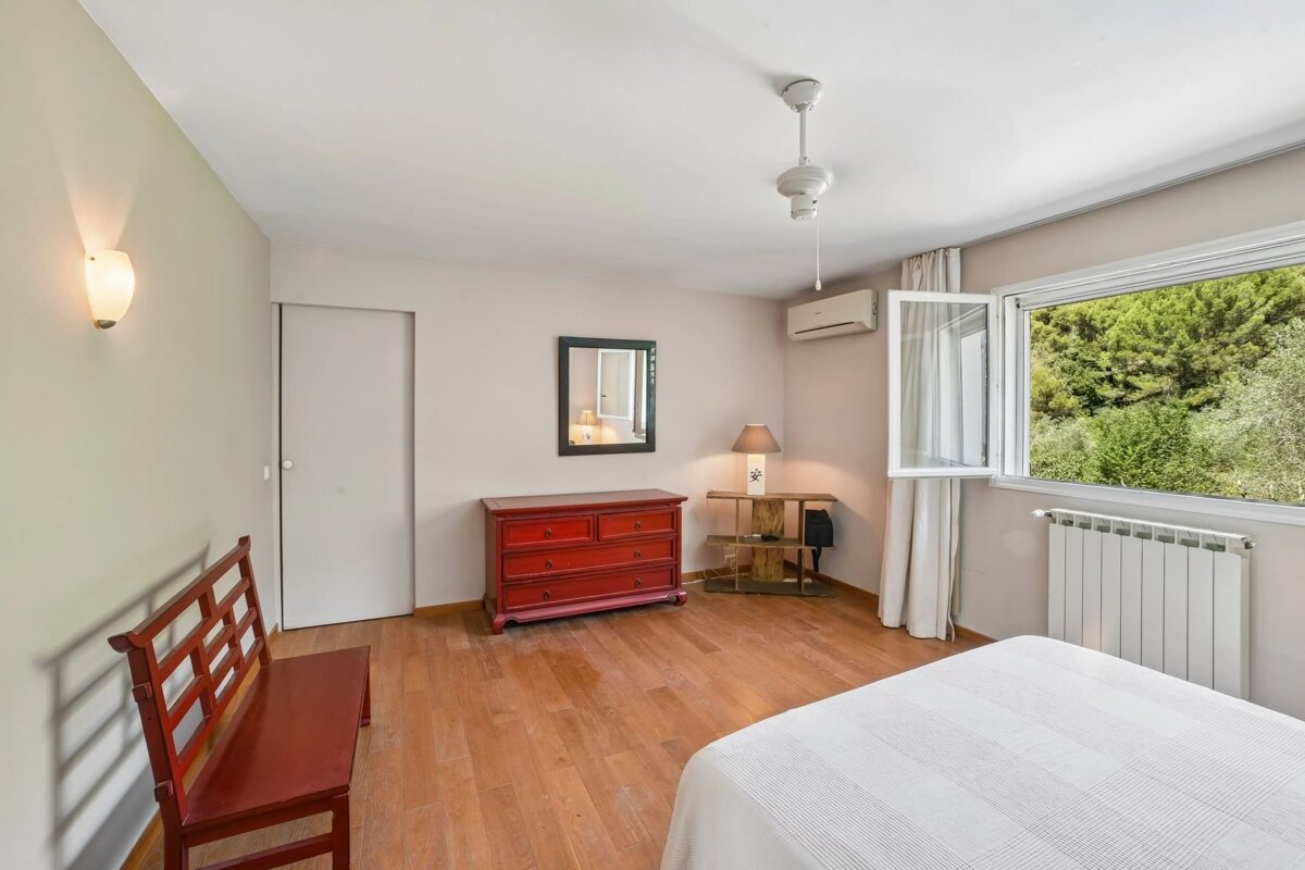Bright bedroom featuring wooden floors, a red dresser, a wooden bench, and an open window with views of lush green trees.