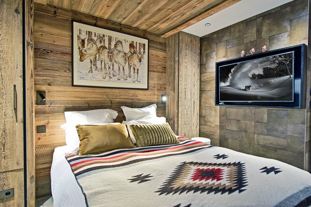 Cozy, rustic bedroom with wood walls, reindeer art, a patterned blanket, and a TV showing a snowboarder.