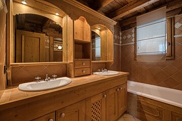 A cozy, rustic wooden bathroom featuring a double vanity with two sinks, mirrors, a bathtub, tiled walls, and exposed wooden ceiling beams.