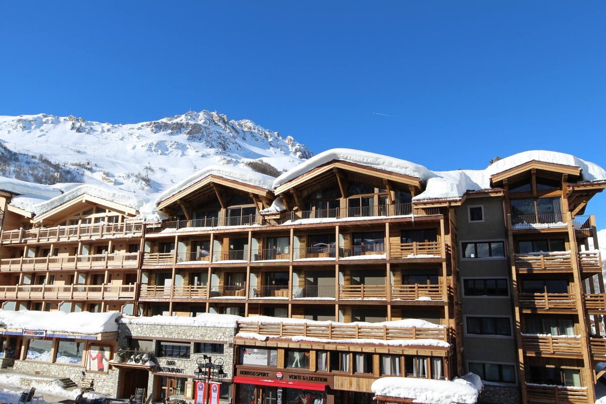 A large wooden ski resort building with snow-covered roofs and balconies stands against majestic snowy mountains under a clear blue sky.