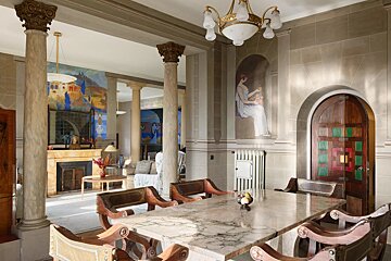 An ornate, neoclassical room with grand columns, a marble dining table, vibrant murals, an elegant chandelier, and a unique arched wooden door.