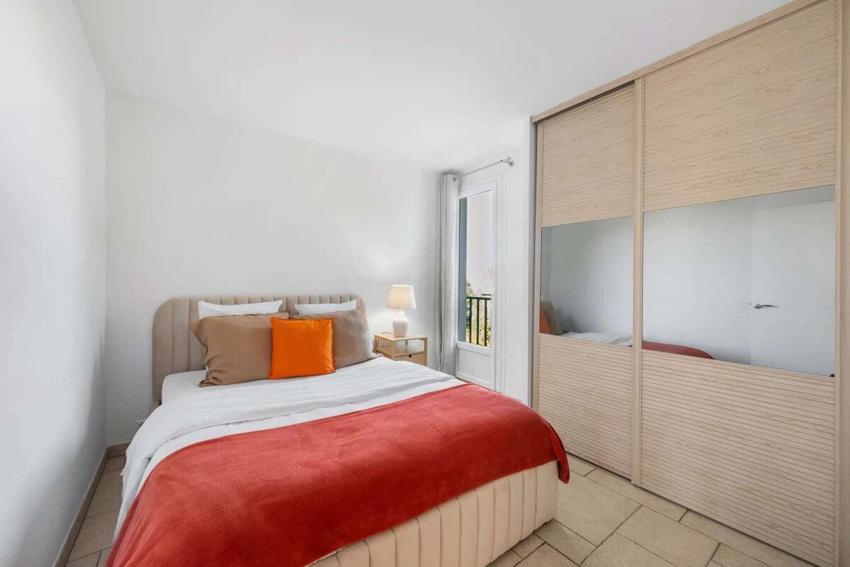 Bright, modern bedroom with a bed, orange blanket, and pillows. Features a nightstand with a lamp, mirrored sliding closet, and balcony view.