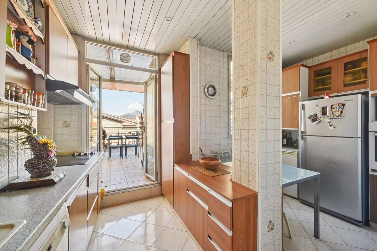 A bright kitchen with tiled floors and wooden cabinets, leading out to a sunny balcony with outdoor seating and a view.