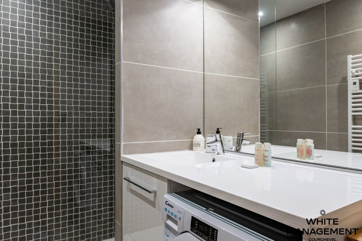 Sleek, modern bathroom featuring a dark mosaic shower, a white vanity with sink, large mirror, and an integrated washing machine. Beige tiles complete the look.