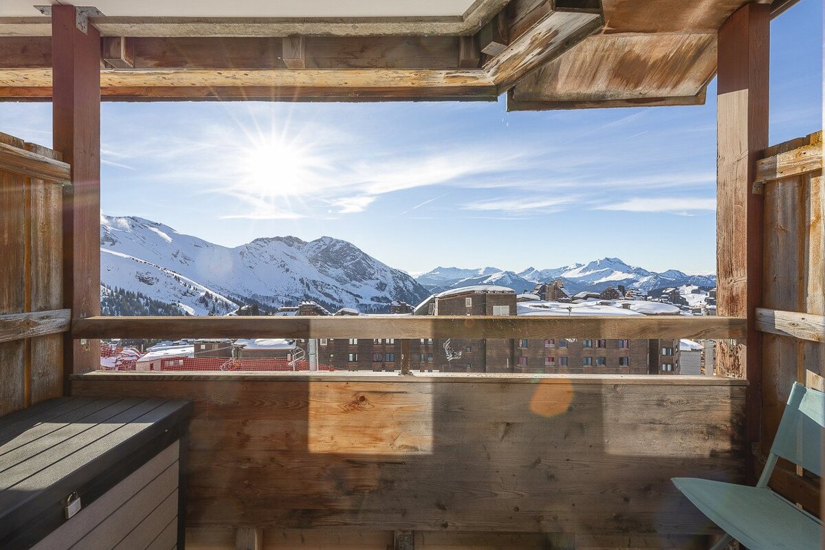 A wooden balcony overlooks sunny, snow-covered mountains and a ski resort under a bright blue sky.