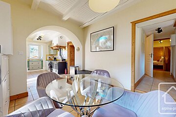 Dining area with glass table & plush chairs, leading to a rustic kitchen via an arch and a bedroom via a hallway, under exposed beams.