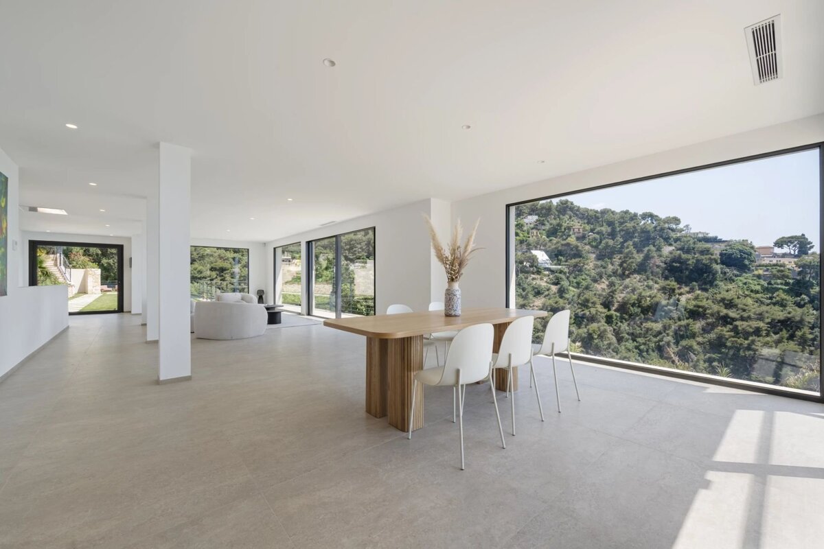 Bright minimalist dining area with a wooden table, white chairs, and a large window showcasing scenic hillside views.