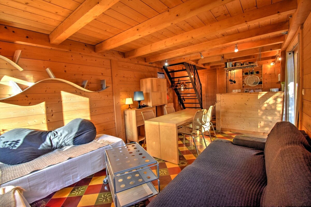Cozy interior of a wooden cabin with a sofa, bed, dining set, patterned tile floor, and stairs leading to a loft.