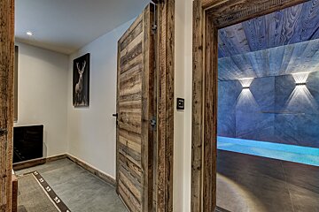 A rustic wooden hallway with a deer picture opens to a modern indoor pool area featuring blue water, unique wall lighting, and a wooden ceiling.