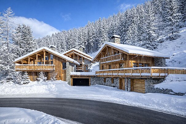 Three rustic wooden chalets in a snow-covered mountain setting, backed by a dense pine forest under a clear blue sky.