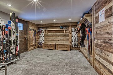 A rustic wooden ski room featuring storage racks, benches, skis, and gear with stone-tile flooring.