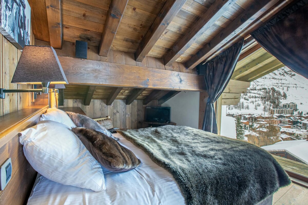 Cozy bedroom with wooden beams and panels, a furry bed, and a large window showcasing a snowy mountain village.