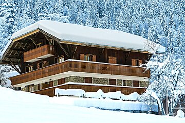 A snowy house with a balcony is surrounded by snow covered trees