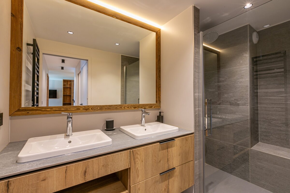 Modern bathroom featuring a wooden double vanity, a large framed mirror, and a walk-in shower with grey tiles.
