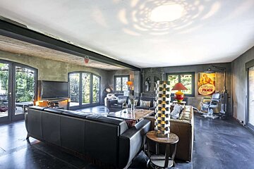 A spacious, dark-toned living room with multiple sofas, arched glass doors, eclectic decor including a barber chair, and unique lighting creating patterns.