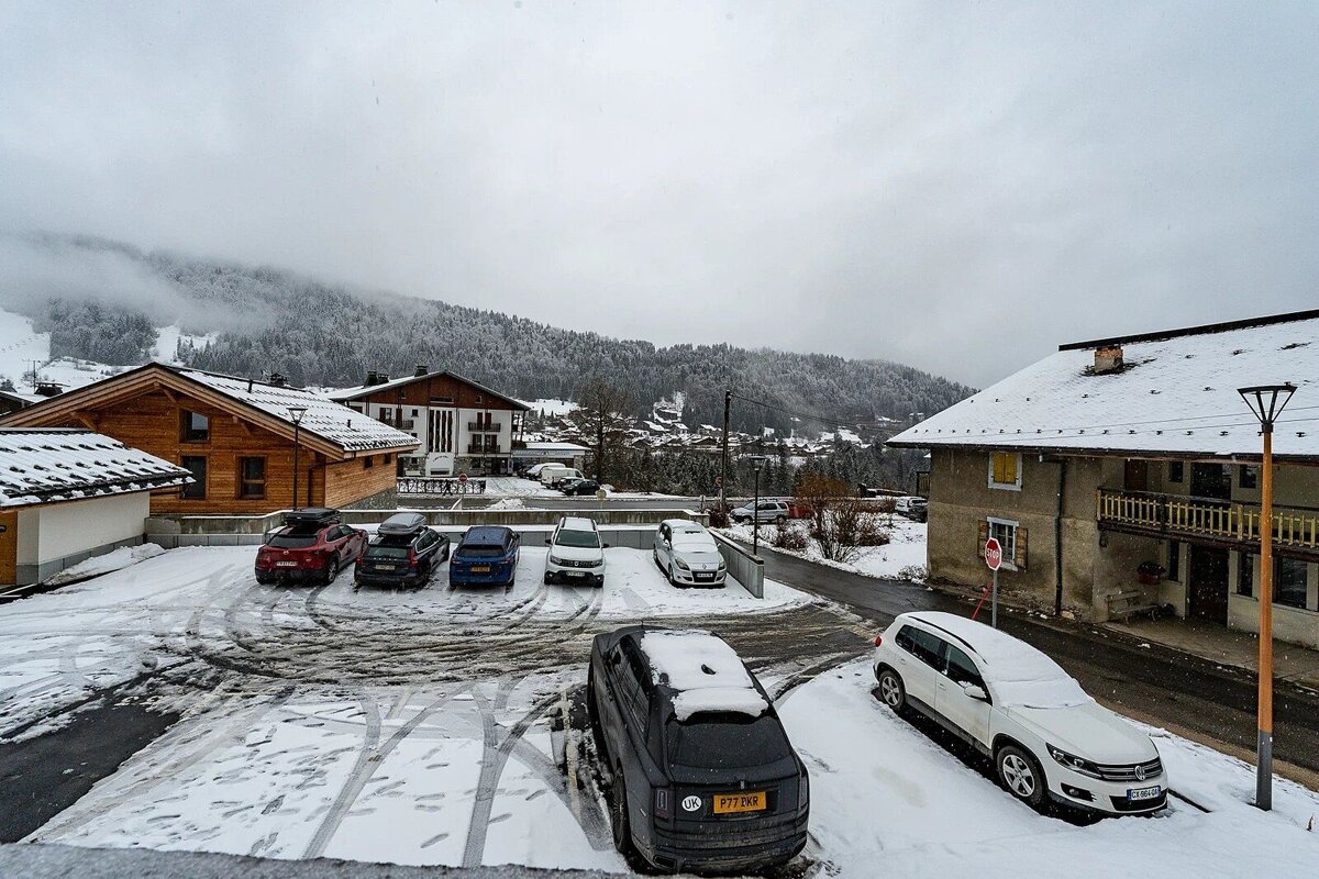 A snow-covered alpine village on an overcast winter day. Cars are parked in a snowy lot, surrounded by chalets and forested mountains.