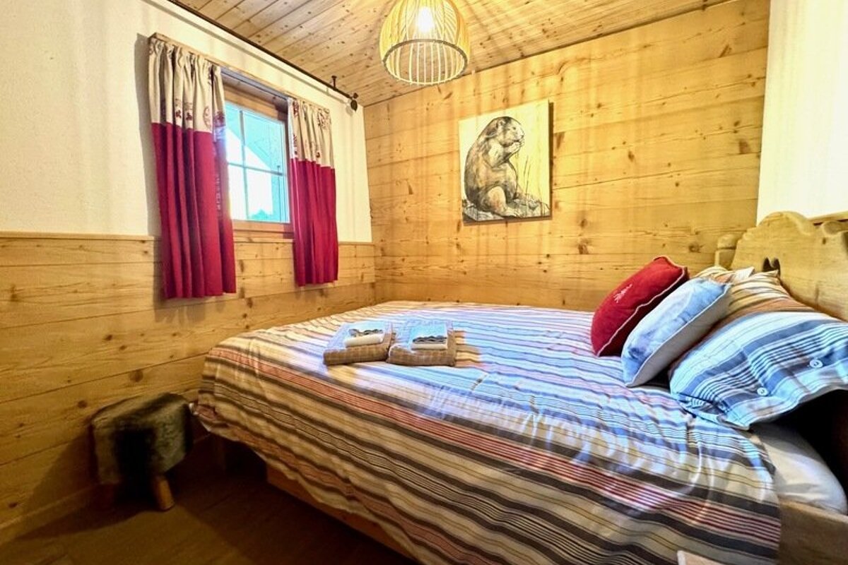 Cozy cabin bedroom with wood-paneled walls, a bed with striped bedding, red curtains, and a marmot art piece on the wall.