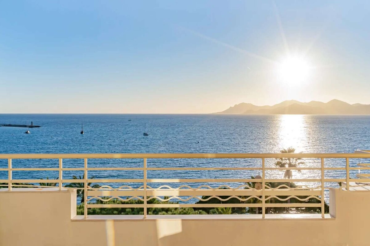 Golden hour over a calm blue ocean, viewed from a balcony. Distant mountains, boats, and a brilliant sun reflecting on the water create a serene scene.