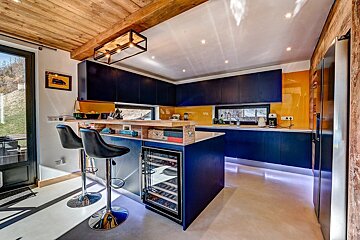 A modern kitchen with navy blue cabinets, yellow backsplash, and wooden ceiling. It features a bar island with stools and a built-in wine cooler.